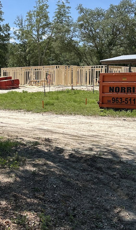 Modern home under construction with exposed framing, red metal staircase, temporary fencing, dirt lot, and scattered building materials; grassy area and trees in background.
