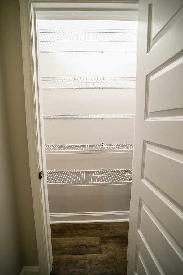 Open white closet door revealing wood floor with white baseboard, white wire shelf, and metal grid shelving against plaster walls