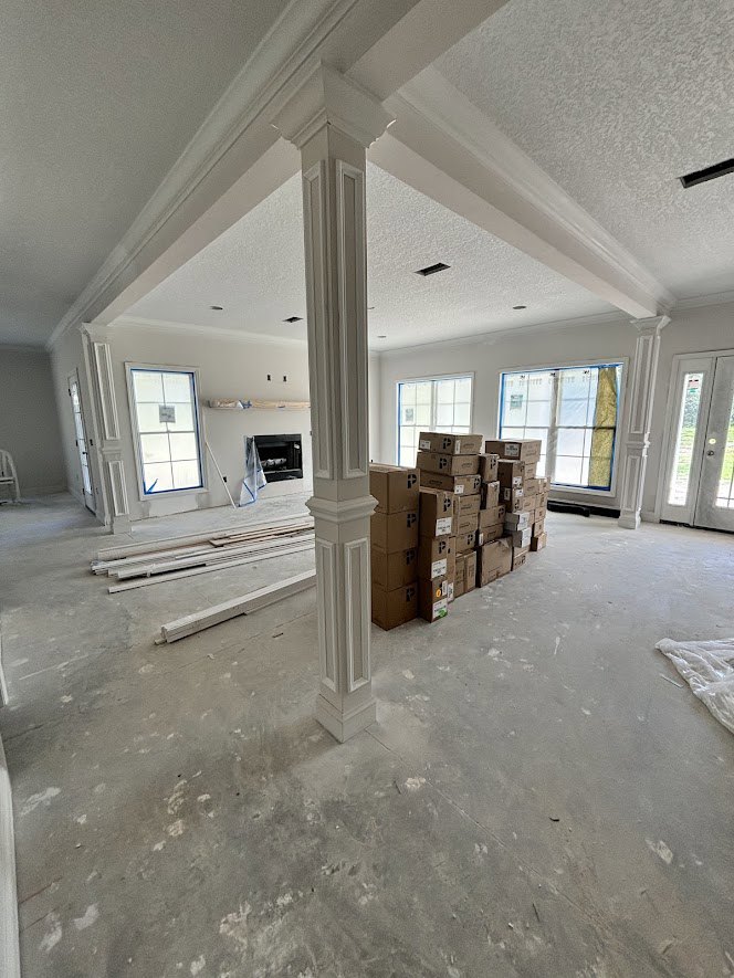 Spacious room with white plaster columns, stacked cardboard boxes on hardwood floor, white door with black trim, large window featuring blue border and white sign