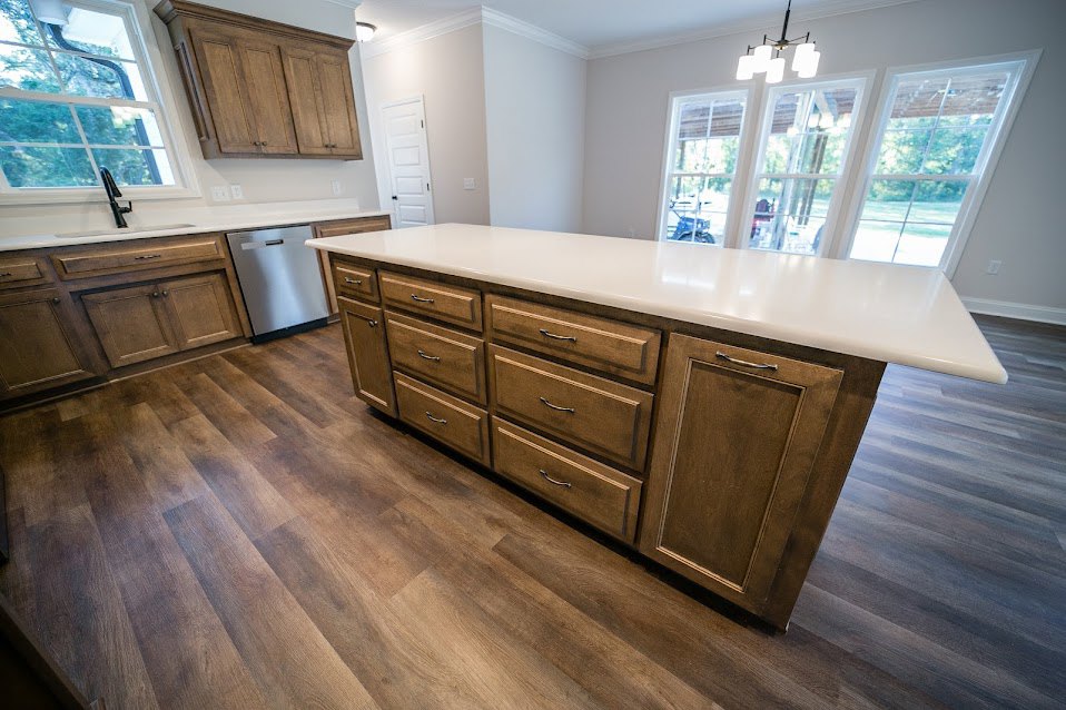 Spacious kitchen featuring a large central island with white countertop, wooden cabinetry, stainless steel dishwasher, multiple drawers, and a four-light pendant fixture above