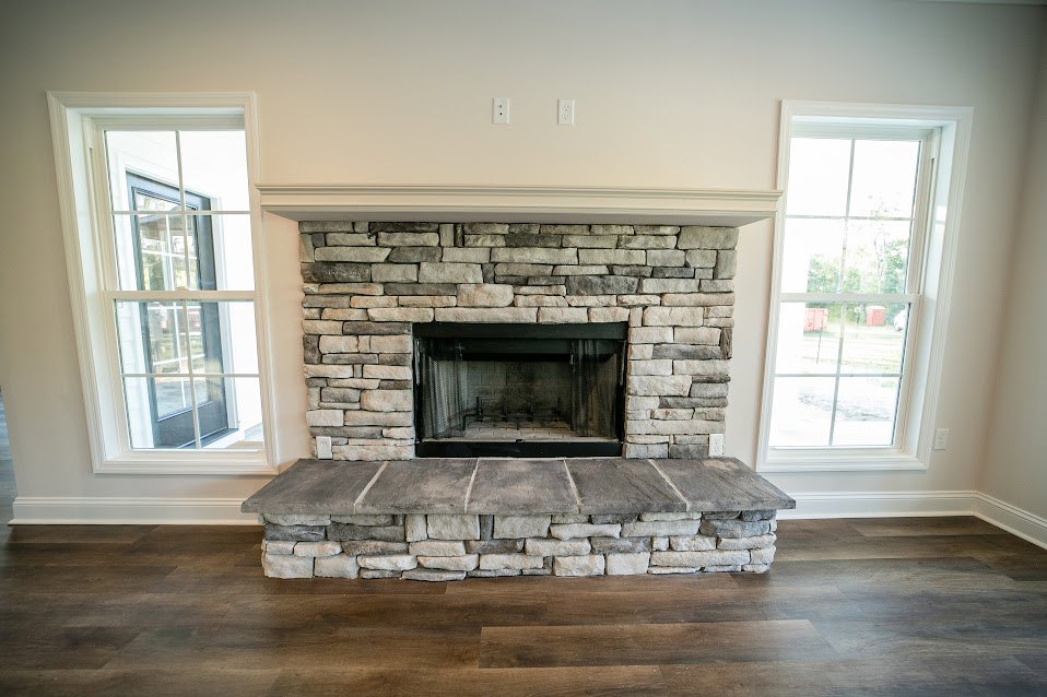 Black-framed fireplace set in a stone wall with stone mantel, white-trimmed window offering outdoor view, hardwood floor, and fire screen in a modern living room.