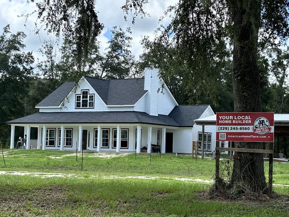 White house with black roof and multiple windows, red real estate sign with white text and image in front yard, tree trunk surrounded by fence, grassy lawn and blue sky.