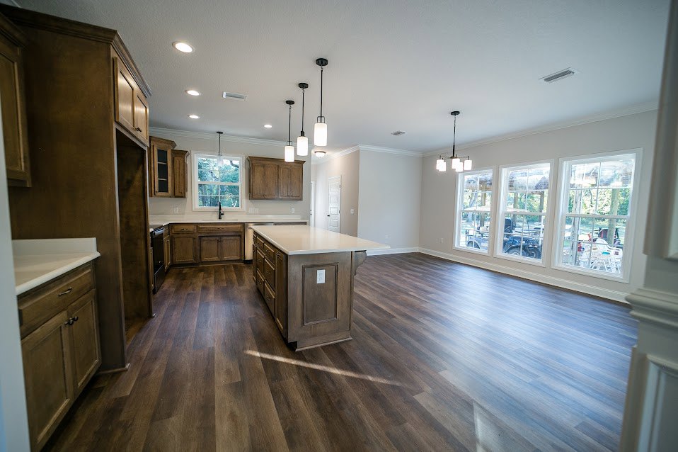 Kitchen with wood flooring, large island featuring a white countertop, pendant light fixture above, wooden cabinets with under-cabinet lighting, row of windows offering exterior