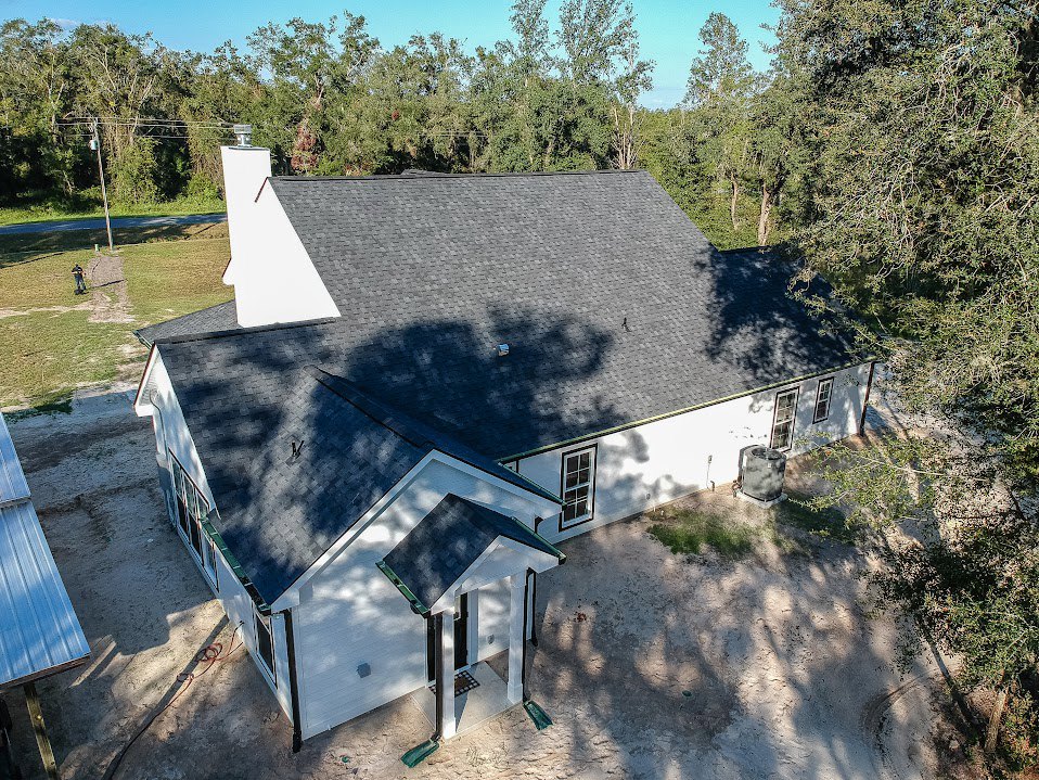 Modern home with black metal roof, light gray siding, large windows, and paved driveway, surrounded by mature trees and green lawn
