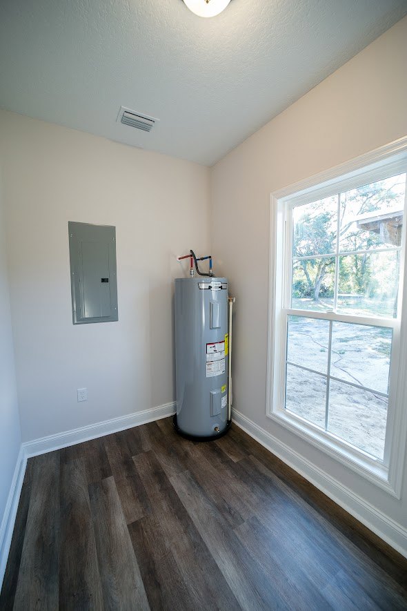 Grey cylindrical water heater with yellow label stands against plaster wall in room with laminate wood flooring and window.