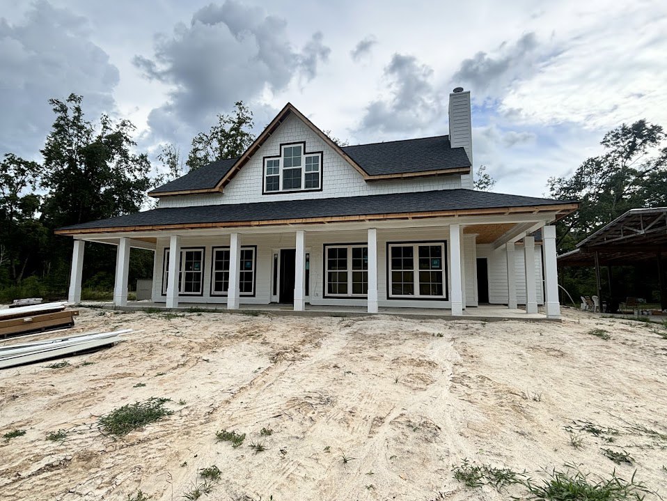 White house with black roof and black door, surrounded by dirt ground with tire tracks and patches of grass, multiple white-framed windows, under cloudy sky.