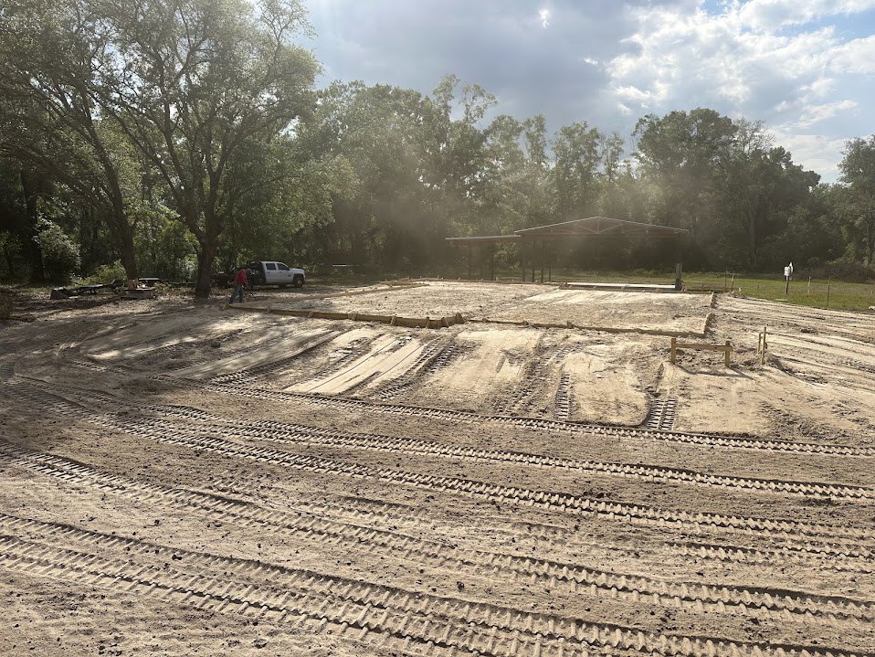 Dirt lot with tire tracks bordered by mature trees under cloudy sky, partially visible roof of building in background
