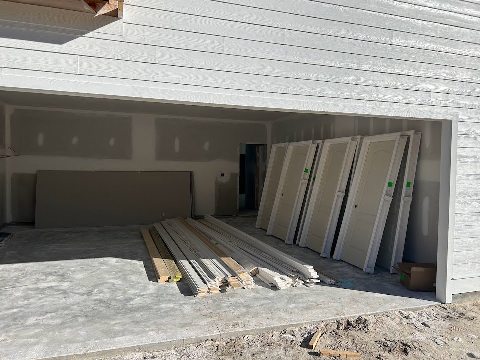 Garage exterior with multiple white doors, wood planks stacked nearby, composite materials on the ground, and a white door featuring a green and white stripe.