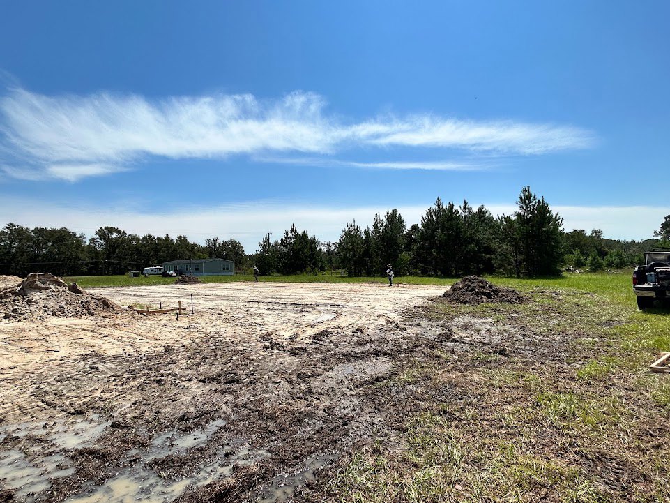 Muddy field with scattered piles of dirt, people standing near a pickup truck, grassy area bordered by trees, custom home visible in the background under a partly cloudy blue sky