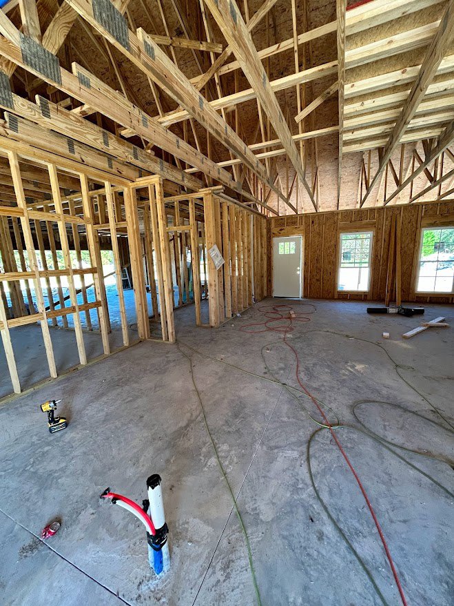 Framed room with exposed wood beams, white door with glass window, concrete floor, visible pipes, and window overlooking green field