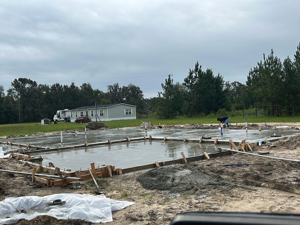 Man spreading cement on a concrete slab foundation beside a house with white trim, cloudy sky and trees in background, knife resting on white sheet nearby
