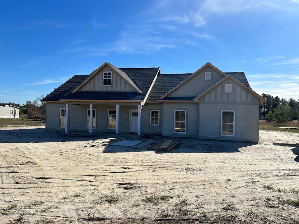 Framed house under construction on dirt lot with white roof, white window frames, and pile of lumber under blue sky