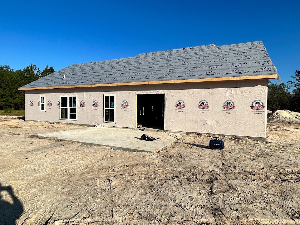 Framed house under construction with exposed plywood walls, white door in dark entryway, multi-pane window, dirt lot with scattered tools, blue sky overhead