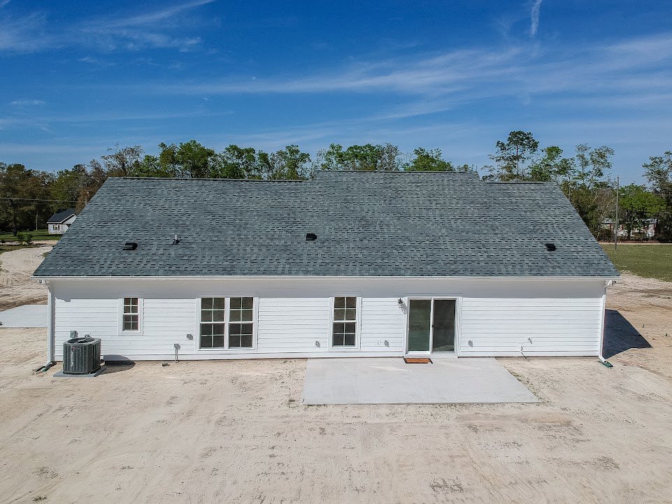 Two-story house with expansive dark roof, white exterior walls, several rectangular windows, and a black air conditioning unit on the side; surrounded by trees and open sky.