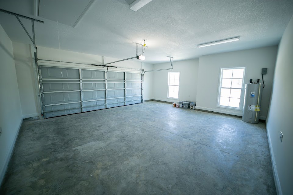 Spacious room featuring polished concrete flooring, white garage door, white-framed window, and water heater against smooth plaster walls