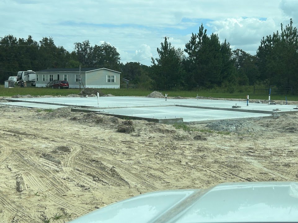 Modern home exterior featuring extensive concrete walls and driveway, white facade, parked car, blue sky with scattered clouds, and surrounding trees.