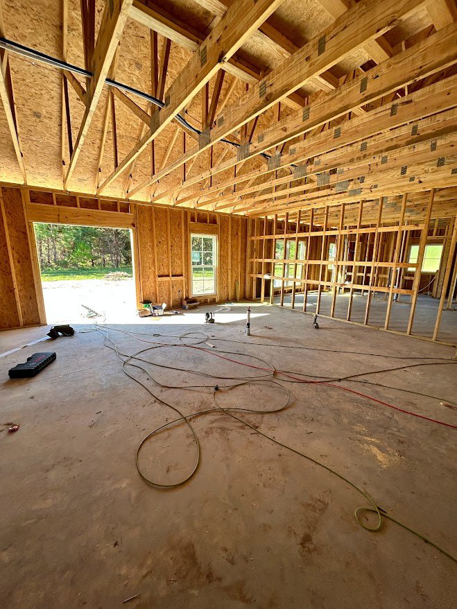 Framed wooden house under construction with exposed beams, large window opening, unfinished floor with electrical wiring, and building insulation visible
