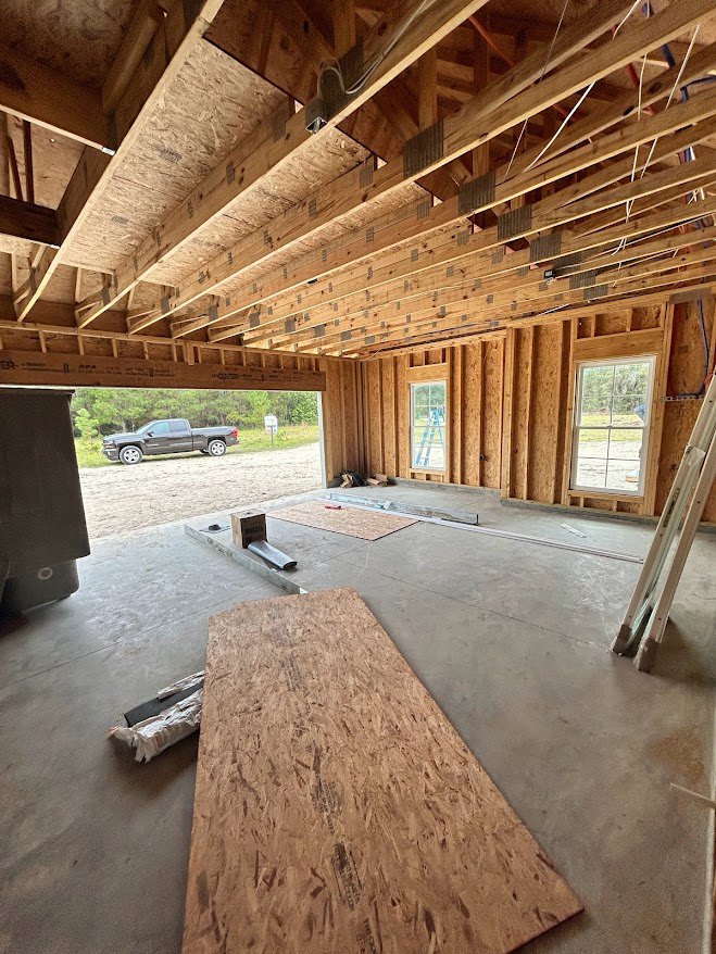 Open room with exposed wood beams and plank ceiling, concrete floor, window overlooking grassy field, garage area visible with parked truck in background