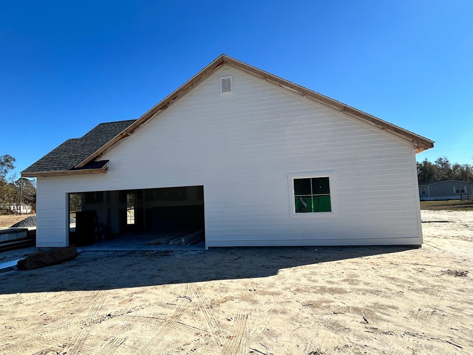 White house under construction with attached garage, exposed roof framing, white siding, visible window with person inside, vent above garage door, dirt ground, trees in background