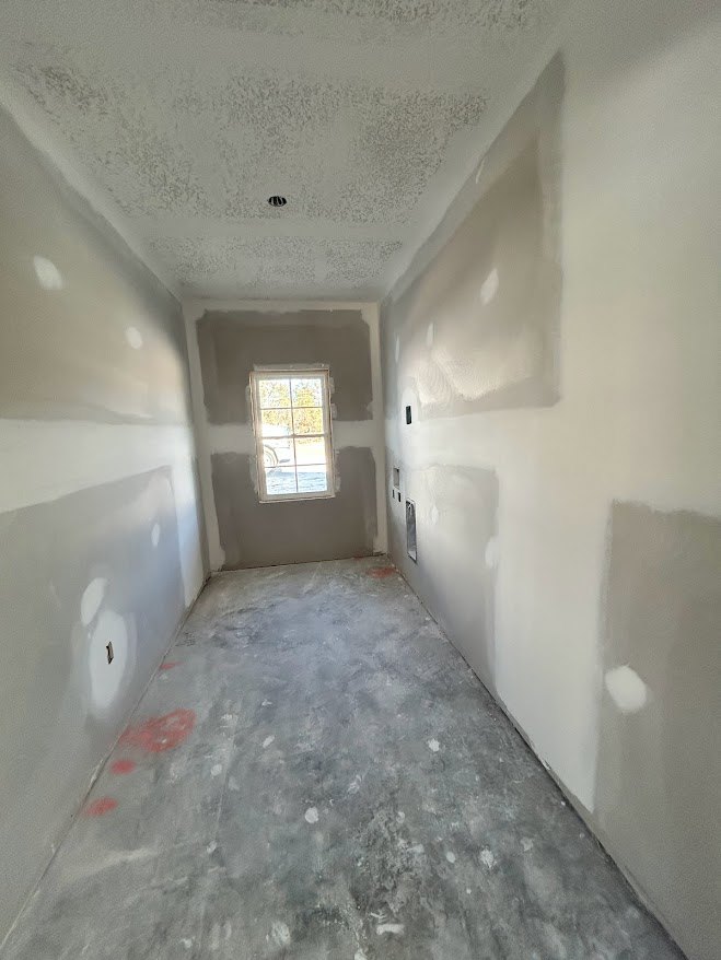 Room with smooth white plaster walls, large window with white frame, exposed concrete floor, ceiling, and visible holes in wall.