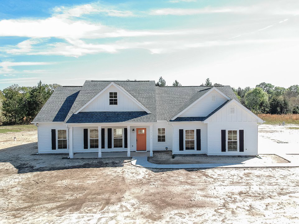 White siding house with black shuttered windows, orange front door, white railing along dirt yard, paved driveway, blue sky with scattered clouds