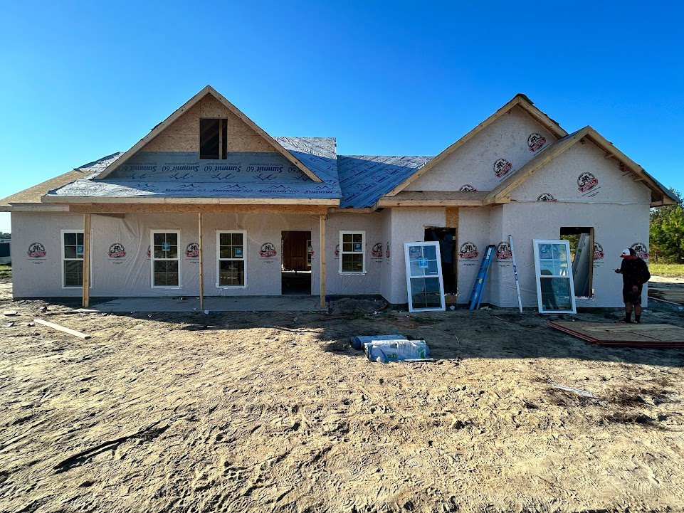Wood-framed house under construction with exposed beams, several ladders leaning against exterior walls, unfinished windows, and clear blue sky overhead
