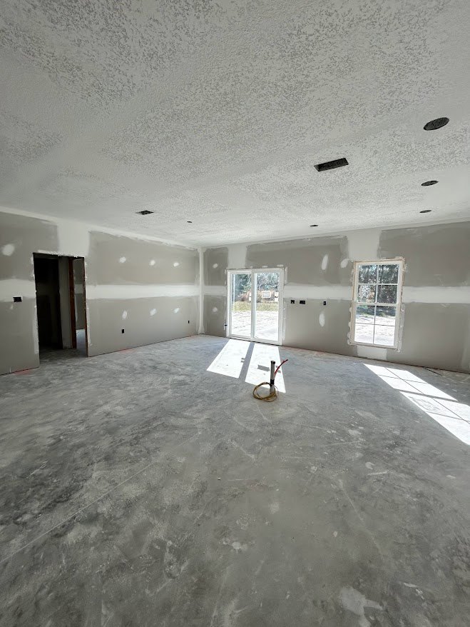 Spacious empty room with plaster walls, large white-framed windows, concrete floor, white door with glass panes, exposed pipe and pole.