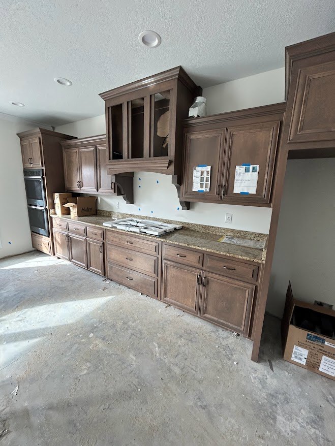 Modern kitchen featuring wood cabinets with glass doors, concrete flooring, stone countertop, stainless steel refrigerator, and a cardboard box with stickers placed near cabinetry.