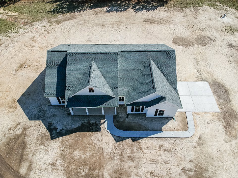 Modern two-story home with light-colored siding, prominent rooflines, concrete driveway, and tree shadows cast across the front yard; person standing on sidewalk near entrance.