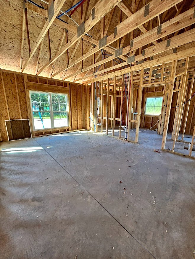 Concrete floor room with exposed wood ceiling beams, white-framed window overlooking greenery, unfinished wooden walls, and construction materials visible.