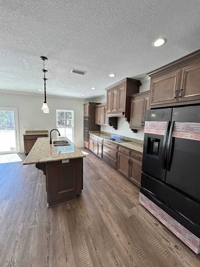 Kitchen with wood flooring, black refrigerator with black handle, central island featuring sink, white-framed window, light cabinetry, and stone countertops