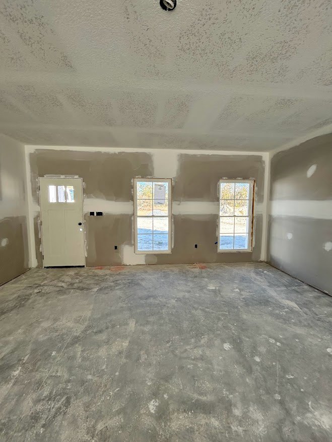 White plaster walls and ceiling, concrete floor, white door with glass panes, two windows showing outdoor view, minimal furnishings