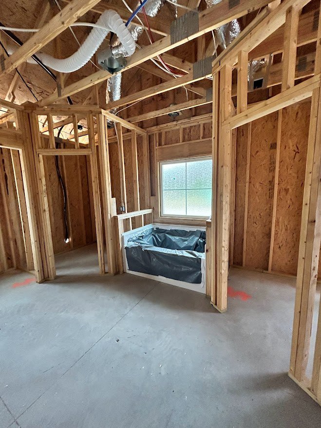 Freestanding square tub with a plastic bag inside, large window, exposed wood beams and planks, unfinished ceiling, hardwood floor, building insulation visible