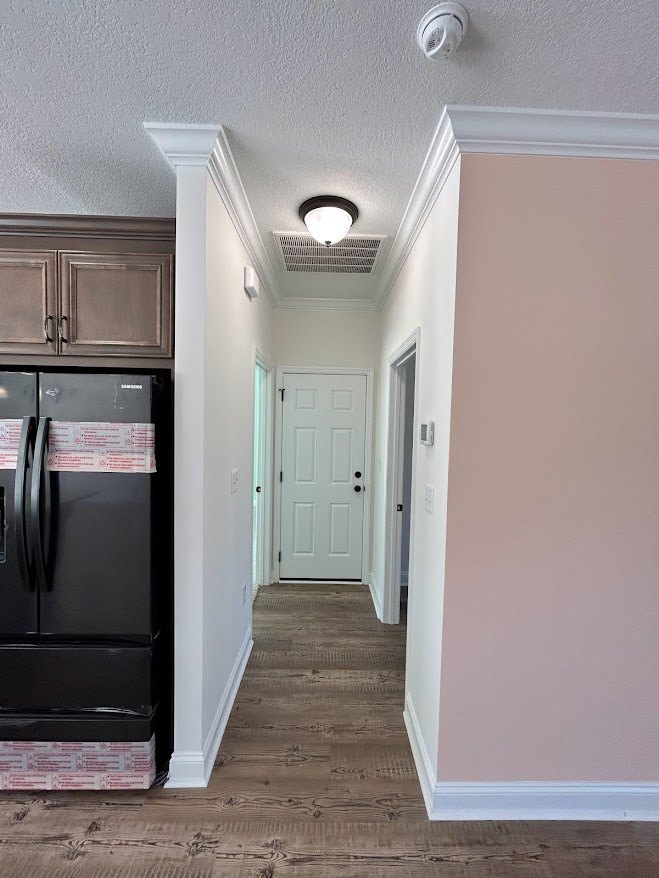 Hallway with light wood flooring, black refrigerator with silver handle, white door with black knobs, open doorway, white ceiling with smoke detector