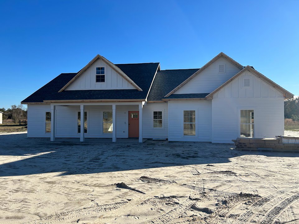 Wood-framed house under construction on sandy ground with tire tracks, red front door, glass window, and pitched roof