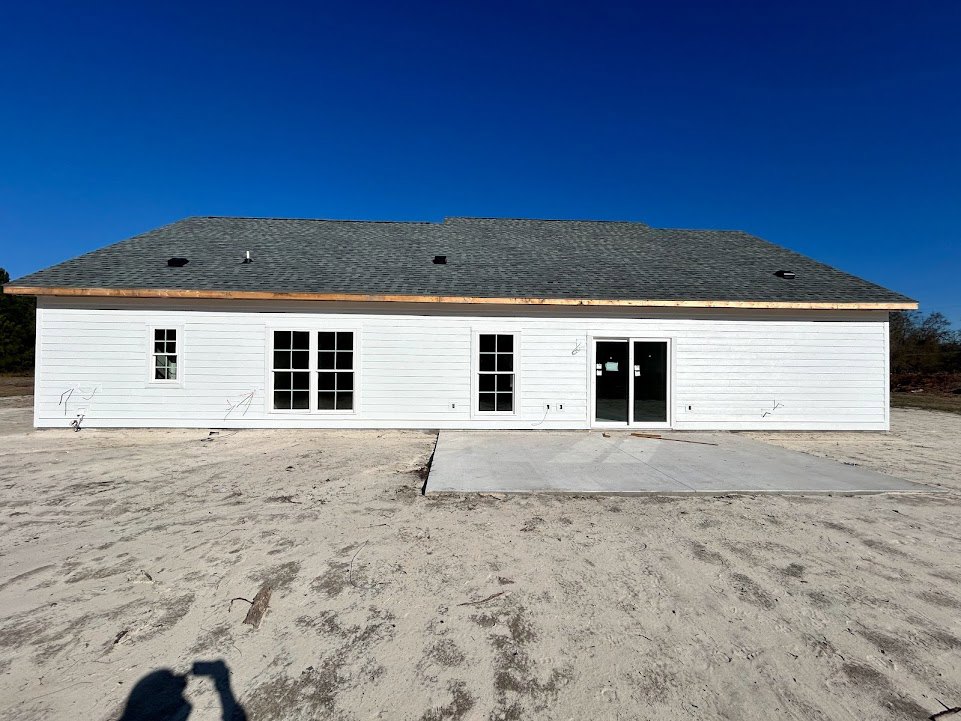 White siding house with black shingle roof, double glass panel doors, grid-style windows, dirt and concrete patio in front