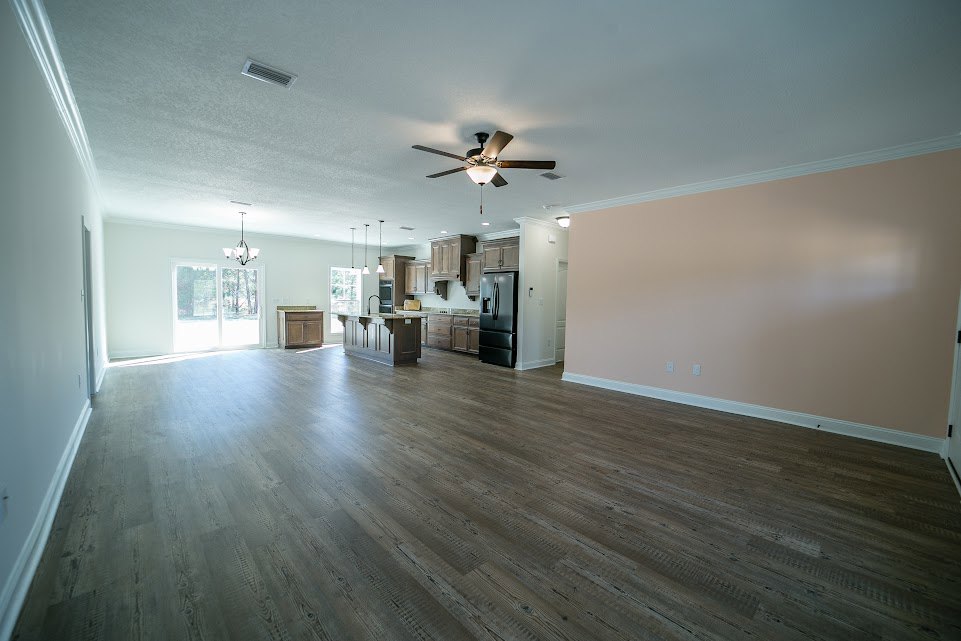 Spacious room with hardwood flooring, ceiling fan with light fixture, pink accent wall, kitchen counter, and sunlight streaming through a window