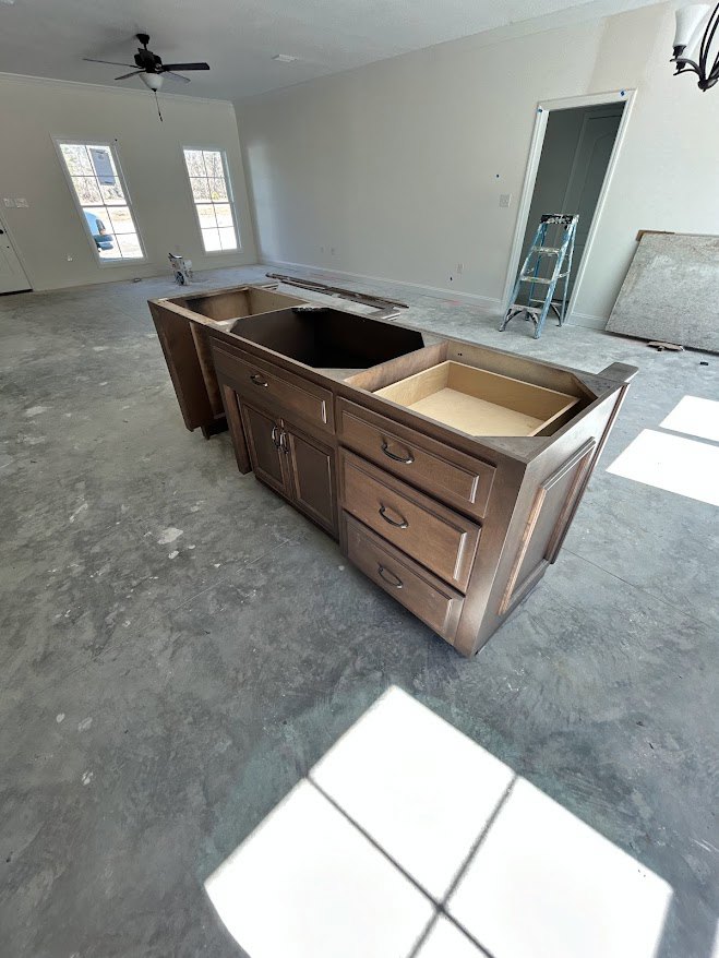 Large kitchen island with multiple drawers, light wood cabinetry, neutral flooring, ceiling fan with light fixture, ladder positioned in doorway, white square object on island