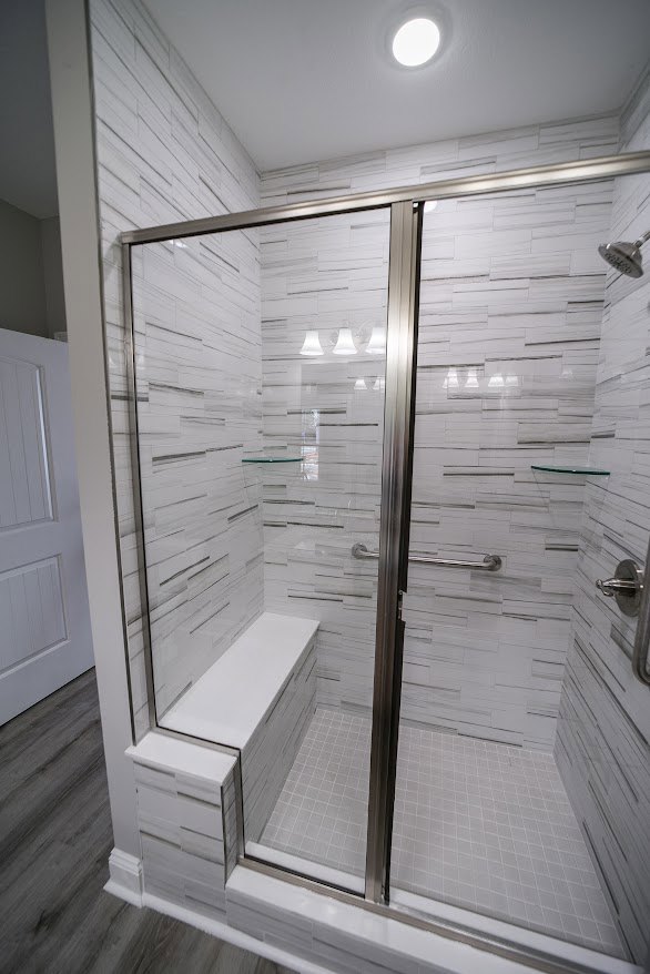 Walk-in shower with clear glass door, white tile walls, chrome fixtures, adjacent white paneled door, dark wood flooring with white baseboards, and white countertop.