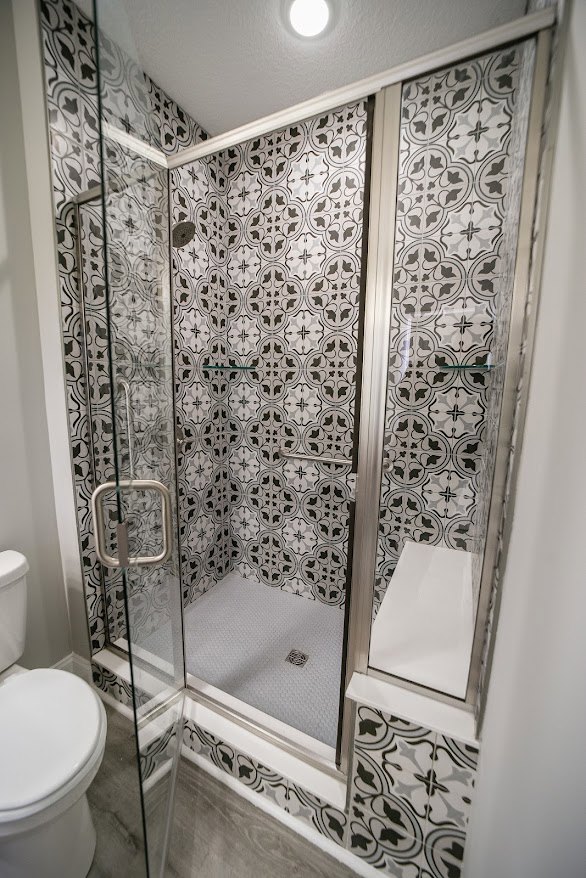 Walk-in shower with clear glass door, white tile walls, adjacent white toilet with closed lid, large wall mirror, chrome fixtures, and light-colored flooring