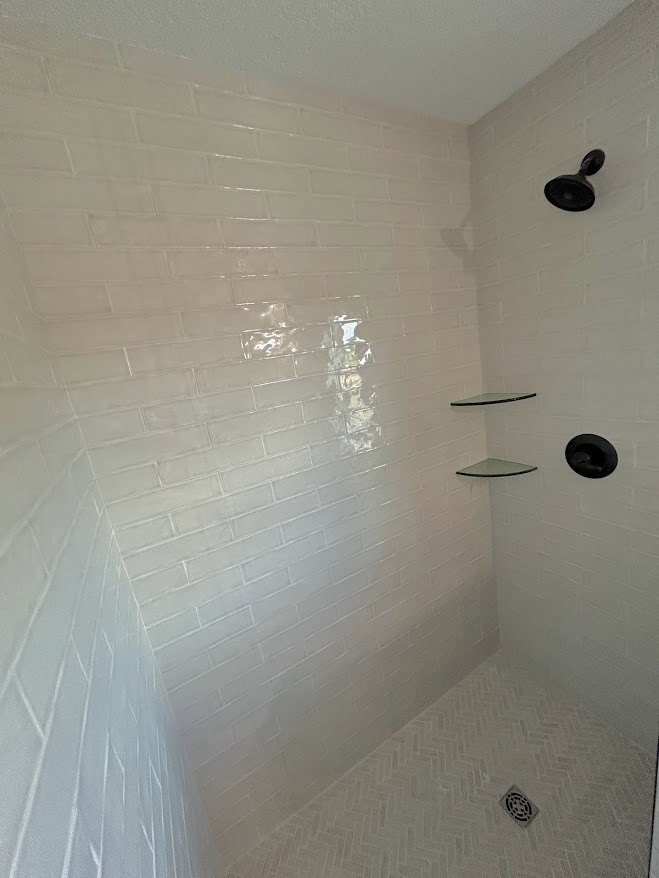 Walk-in shower with built-in shelves, white brick tile walls, gray tile floor, ceiling-mounted black showerhead, circular drain, and plaster ceiling