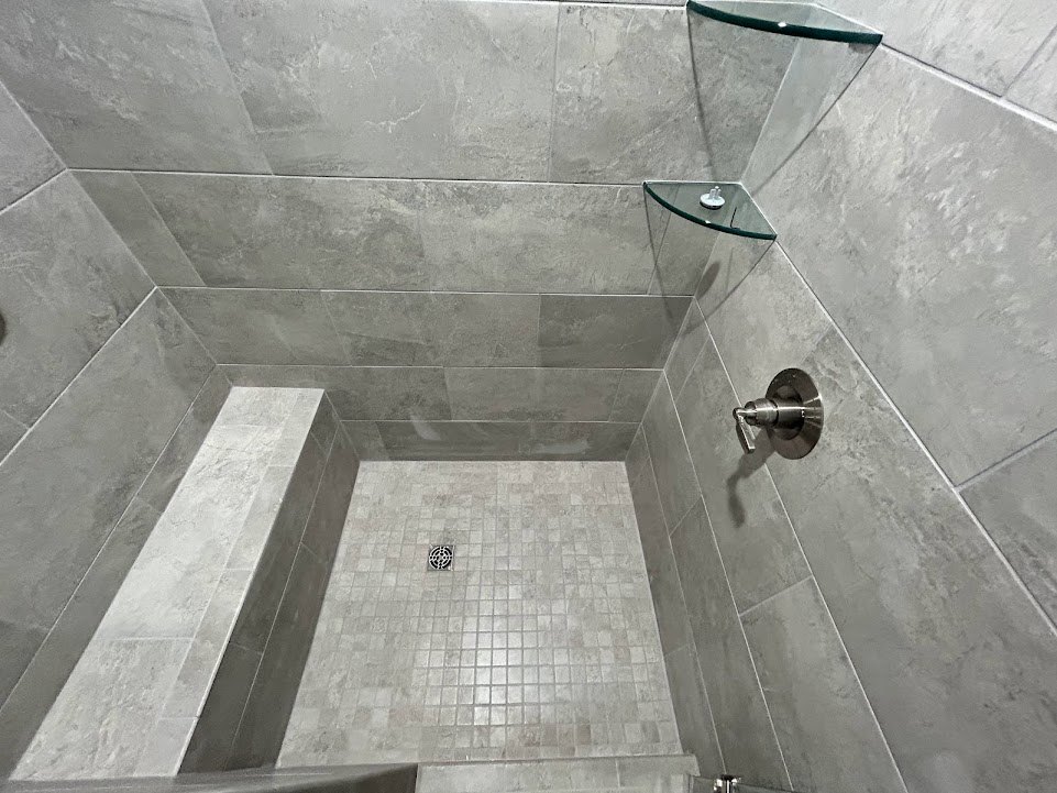 Marble shower with glass shelf, chrome handle, and linear drain; white soap dish rests on transparent shelf