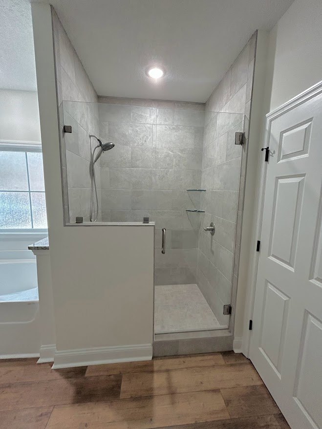 Bathroom featuring a glass shower enclosure, marble tile floor, white walls, wood flooring with white baseboard, and a close-up of a door adjacent to the shower.