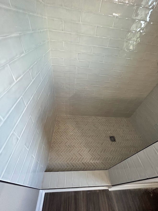 Walk-in shower with white tile walls, chrome shower head, and gray tile floor adjacent to wood flooring