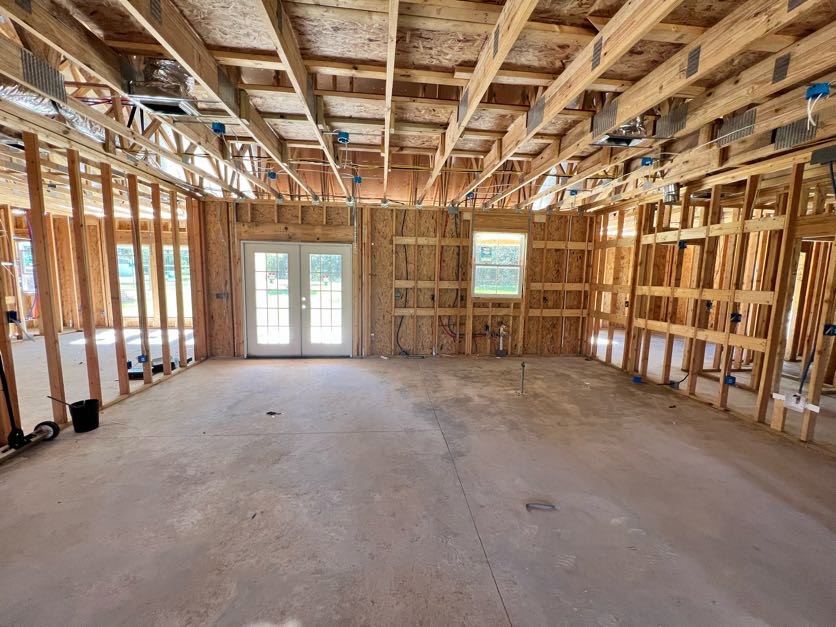 Spacious room with concrete floor featuring central cutout, double glass-paneled doors, large window displaying a sign, exposed wooden ceiling beams with blue tape, and