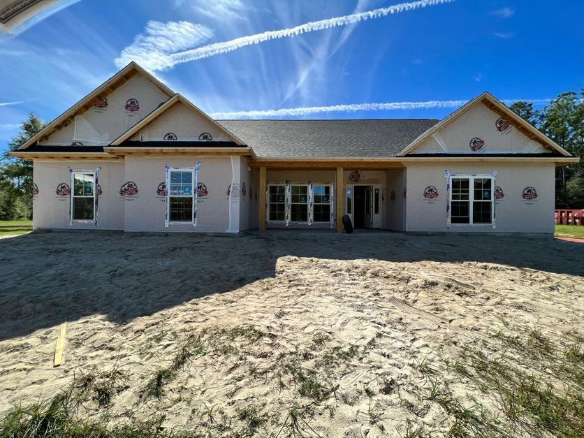 Two-story house under construction with sandy soil in front, white-framed windows with multiple panes, unfinished exterior walls, and blue sky overhead