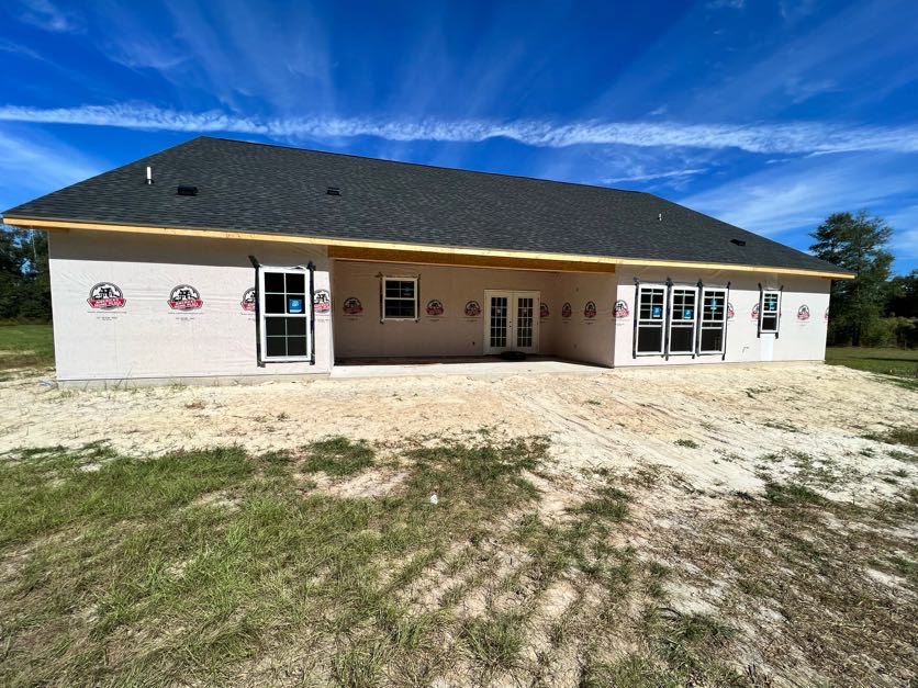 Modern home exterior with light stucco walls, double glass doors, multiple windows featuring blue stickers, dirt yard with a small grass patch, cloudy sky overhead