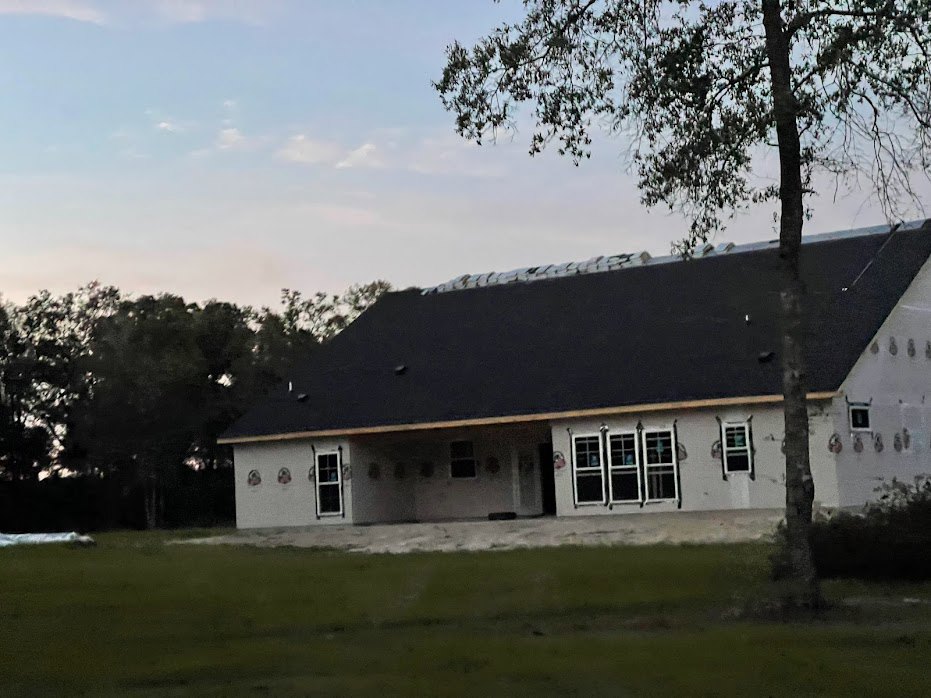 White house with black roof, large windows, manicured lawn, mature trees, and cloudy sky in the background