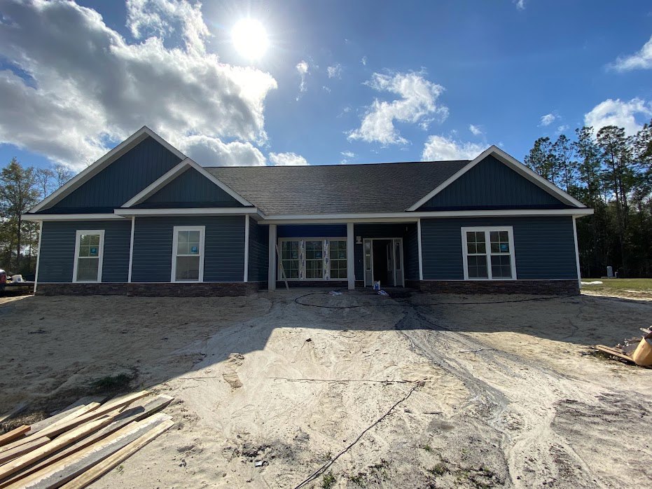 Two-story house under construction with blue roof, dirt driveway, pile of wood planks in front, white-framed windows, one window displaying a blue sign, and a ladder leaning