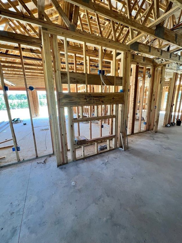Exposed wood framing and beams inside a house under construction, unfinished walls with window openings, plywood floor, and visible ceiling structure